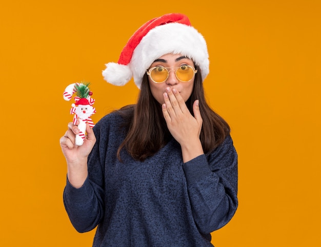 Удивленная молодая кавказская девушка в солнцезащитных очках в шляпе санта-клауса держит конфету и кладет руку на рот, изолированную на оранжевой стене с копией пространства