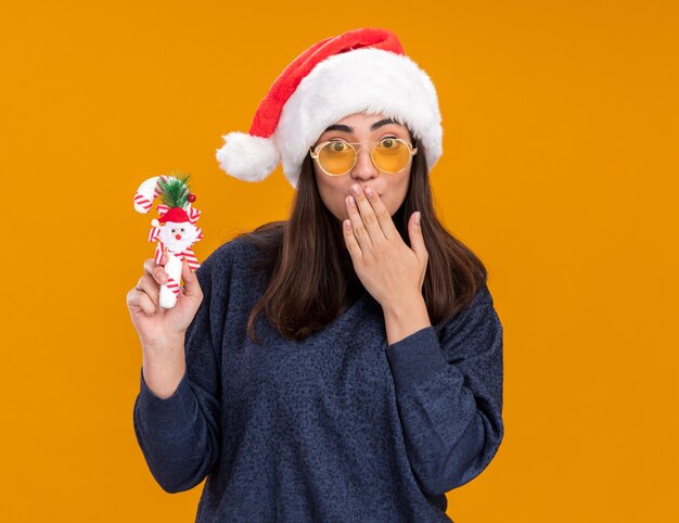 Surprised young caucasian girl in sun glasses with santa hat holds candy cane and puts hand on mouth isolated on orange wall with copy space