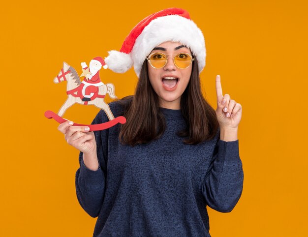 Surprised young caucasian girl in sun glasses with santa hat holds santa on rocking horse decoration and points up isolated on orange wall with copy space