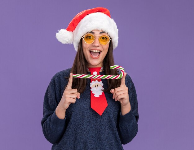 Surprised young caucasian girl in sun glasses with santa hat and santa tie holding candy cane isolated on purple background with copy space