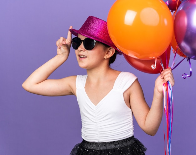 surprised young caucasian girl in sun glasses with violet party hat holding helium balloons and pointing up isolated on purple wall with copy space