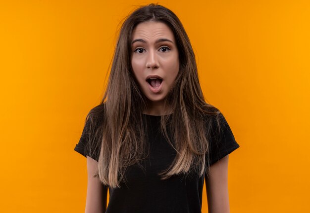 Surprised young caucasian girl wearing black t-shirt looking at camera on isolated orange background