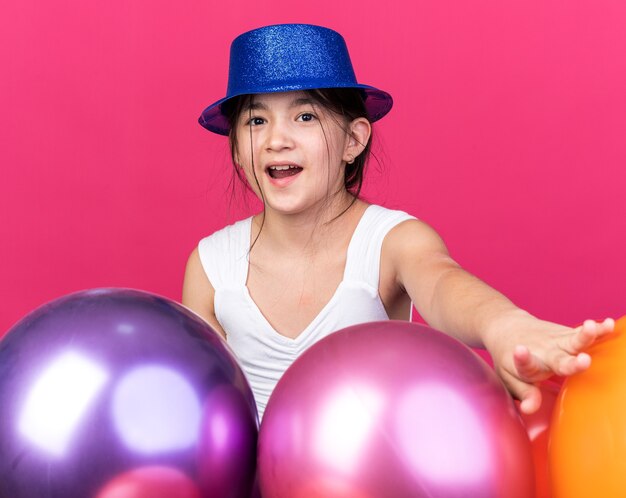 コピースペースでピンクの壁に分離されたヘリウム気球と立っている青いパーティーハットを身に着けている若い白人の女の子を驚かせた