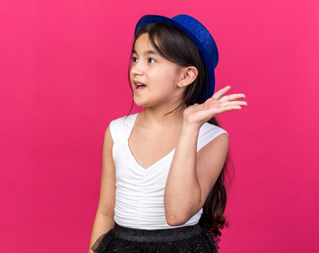 surprised young caucasian girl with blue party hat standing with raised hand looking at side isolated on pink wall with copy space