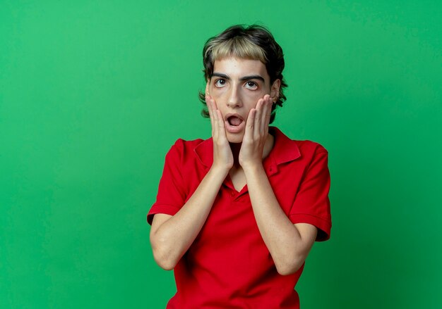 Surprised young caucasian girl with pixie haircut putting hands on face isolated on green background with copy space