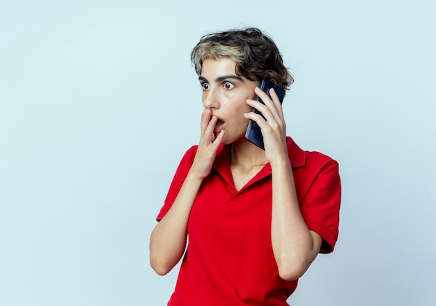 Удивленная молодая кавказская девушка со стрижкой пикси разговаривает по телефону, положив руку на рот, глядя прямо на белом фоне с копией пространства