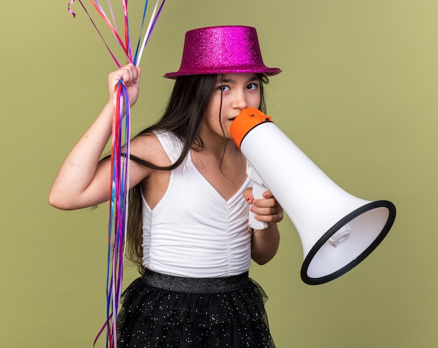 Foto gratuita sorpresa giovane ragazza caucasica con cappello da festa viola che tiene palloncini di elio e altoparlante isolato su parete verde oliva con spazio di copia