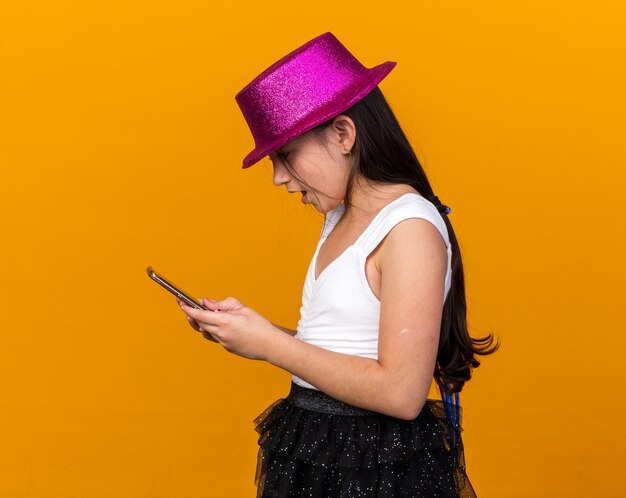 Sorpresa giovane ragazza caucasica con cappello da festa viola che tiene e guarda il telefono isolato sulla parete arancione con spazio di copia copy