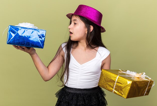 surprised young caucasian girl with purple party hat looking at gift boxes holding on each hand isolated on olive green wall with copy space