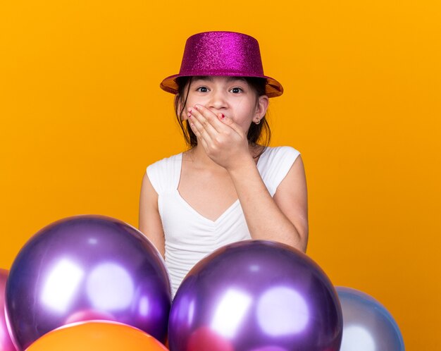 surprised young caucasian girl with purple party hat putting hand on mouth standing with helium balloons isolated on orange wall with copy space