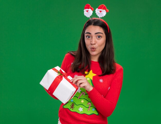 Удивленная молодая кавказская девушка с повязкой на голову санта-клауса держит рождественскую подарочную коробку, изолированную на зеленой стене с копией пространства
