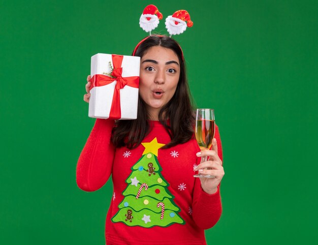 Surprised young caucasian girl with santa headband holds glass of champagne and christmas gift box isolated on green wall with copy space