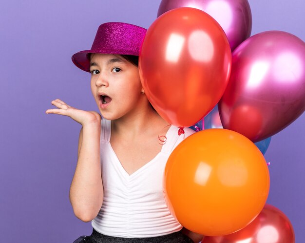 ヘリウム気球を保持し、コピースペースで紫色の壁に隔離された手を開いたままにしておく紫色のパーティハットで驚いた若い白人の女の子