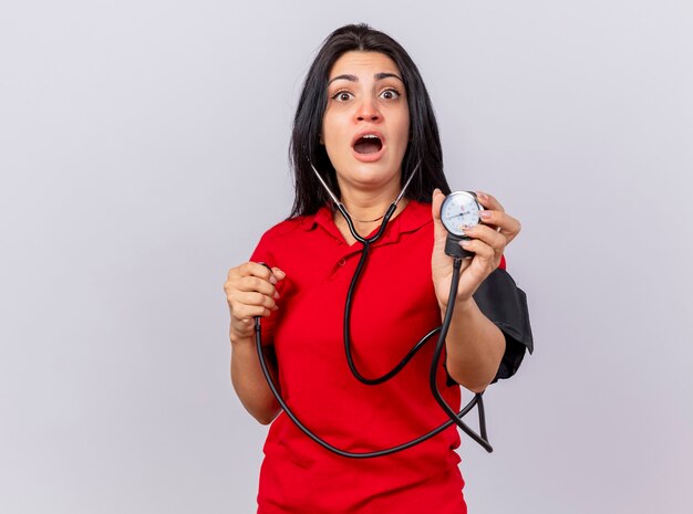 Surprised young caucasian ill girl wearing stethoscope looking at camera measuring her pressure with sphygmomanometer isolated on white background with copy space