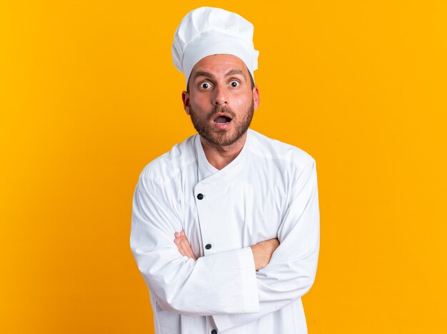 Surprised young caucasian male cook in chef uniform and cap standing with closed posture 