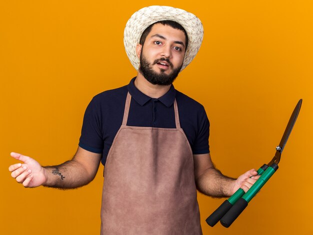 surprised young caucasian male gardener wearing gardening hat holding gardening scissors isolated on orange wall with copy space