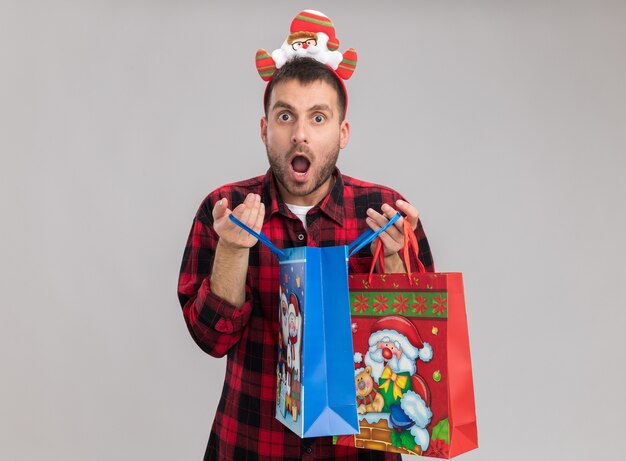 surprised young caucasian man wearing christmas headband holding christmas gift bags opening one looking  isolated on white wall with copy space