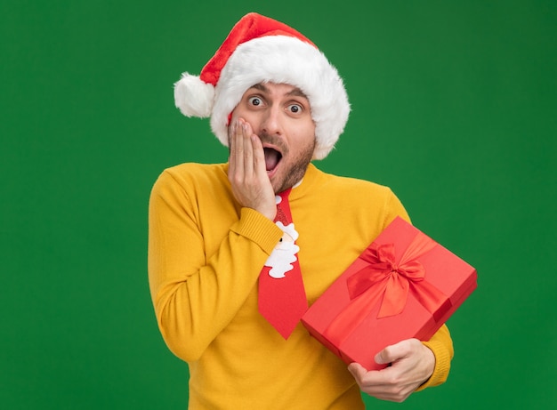 Surprised young caucasian man wearing christmas tie and hat holding gift package looking at camera keeping hand on face isolated on green background with copy space