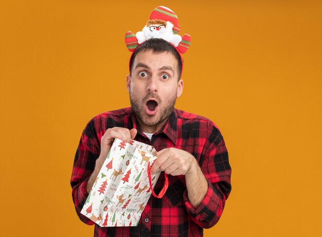 surprised young caucasian man wearing santa claus headband holding christmas gift bag looking  isolated on orange wall with copy space