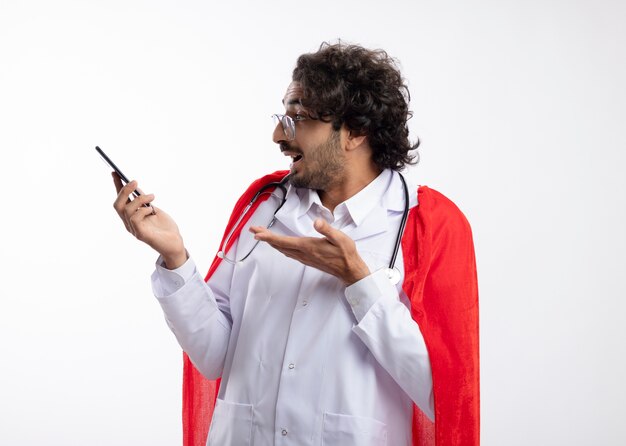 Surprised young caucasian superhero man in optical glasses wearing doctor uniform with red cloak and with stethoscope around neck looks and points at phone 