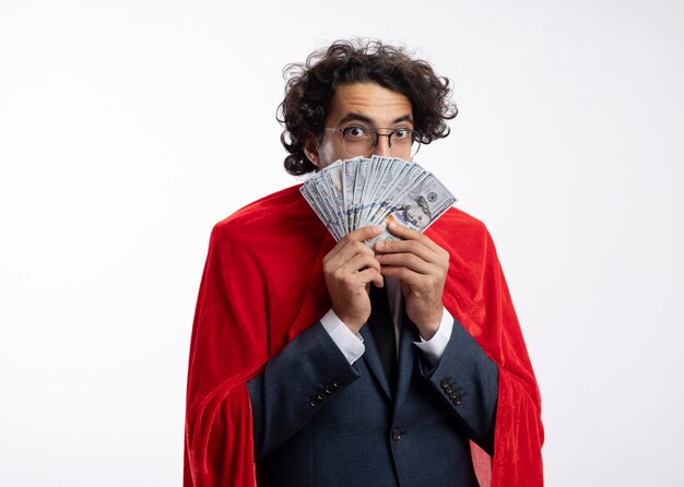 Surprised young caucasian superhero man in optical glasses wearing suit with red cloak holds money 