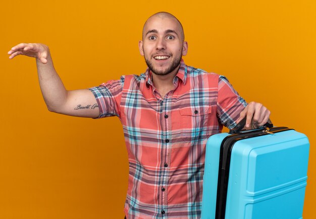 Surprised young caucasian traveler man holding suitcase and keeping hand open isolated on orange wall with copy space