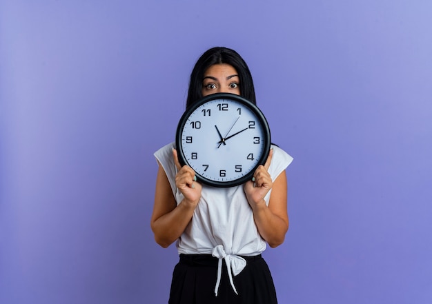 Surprised young caucasian woman holds and looks over clock