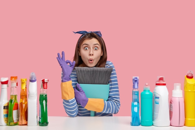 surprised young Caucasian woman makes okay gesture, keeps broom closely, shows everything is ok, sweeps floor