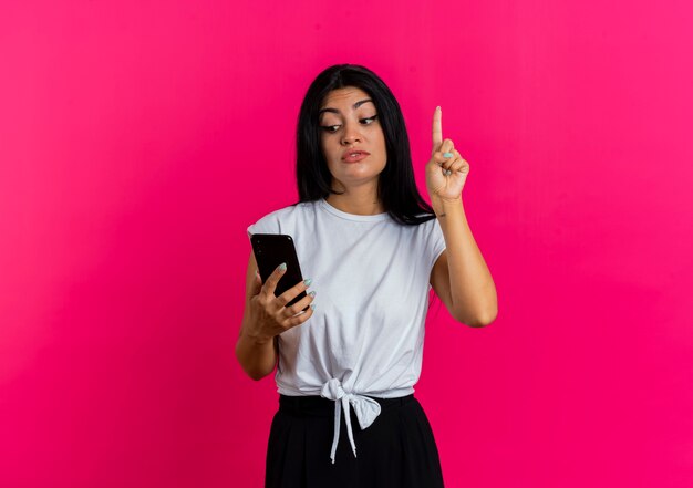 Surprised young caucasian woman points up and looks at phone