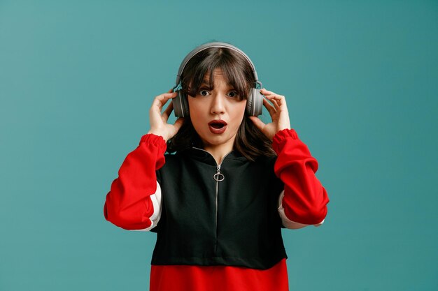 Surprised young caucasian woman wearing and grabbing headphones looking at camera isolated on blue background