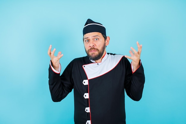 Surprised young chef is looking at camera by raising up his hands on blue background