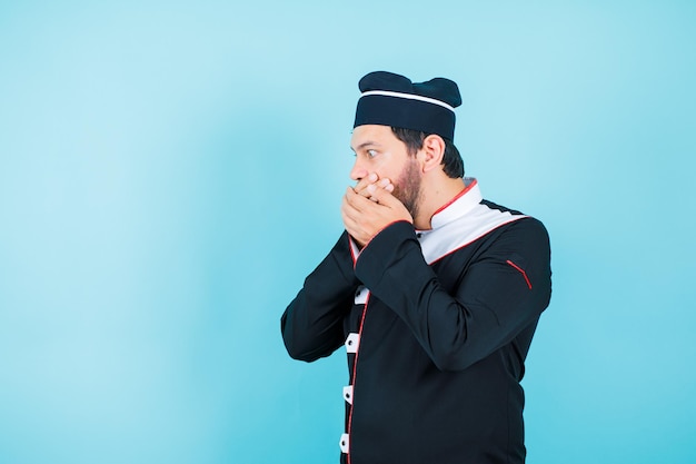 Surprised young chef is looking left side by holding hands on mouth on blue background