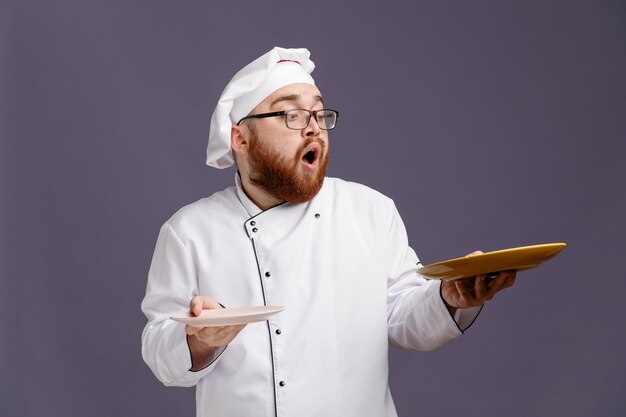 Surprised young chef wearing glasses uniform and cap holding empty plates looking at one isolated on purple background