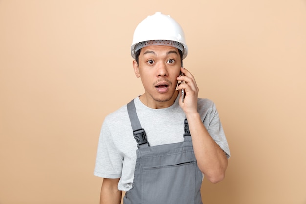 Surprised young construction worker wearing safety helmet and uniform talking on phone 