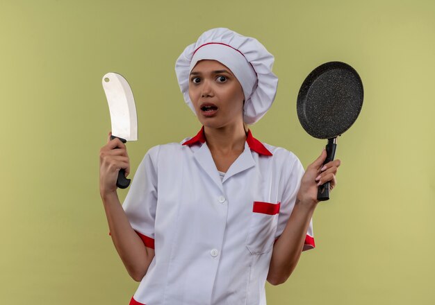 프라이팬과 칼을 들고 요리사 유니폼을 입고 놀란 젊은 요리사 여성