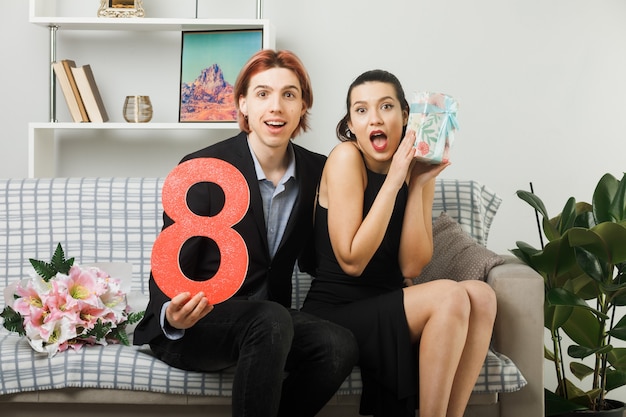 Surprised young couple on happy women day holding number eight with present sitting on sofa in living room