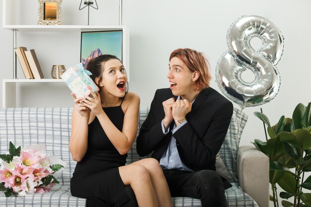 Surprised young couple on happy women day holding present sitting on sofa in living room