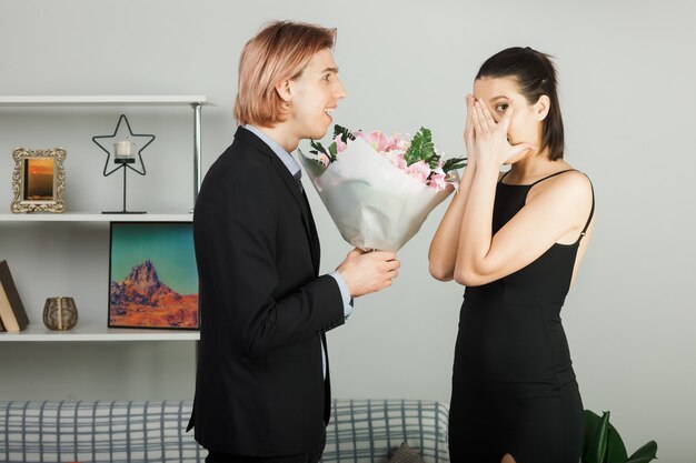 Surprised young couple hugged each other on valentines day with bouquet girl whispers standing in living room