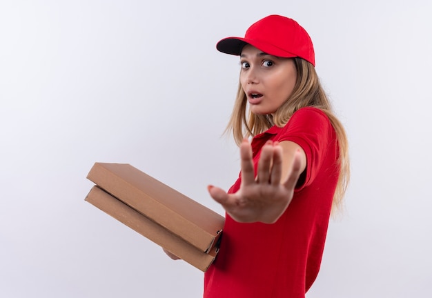 Удивленная молодая доставщица в красной форме и кепке держит коробки для пиццы и показывает жест остановки, изолированный на белом