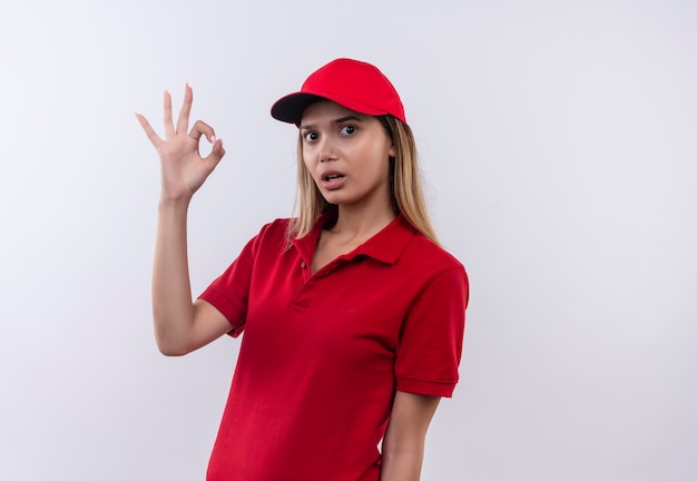 удивленная молодая доставщица в красной униформе и кепке, показывающая хороший жест, изолирована на белой стене