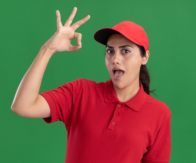 Surprised young delivery girl wearing uniform and cap showing okay gesture isolated on green wall