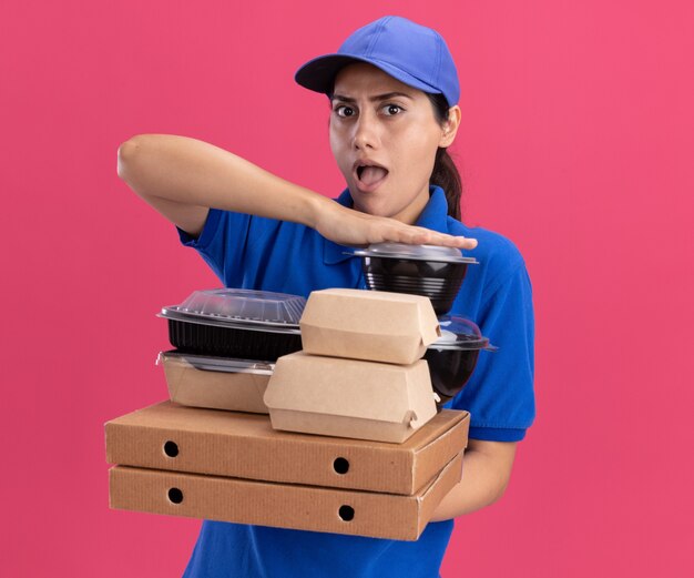 Surprised young delivery girl wearing uniform with cap holding food containers on pizza boxes isolated on pink wall