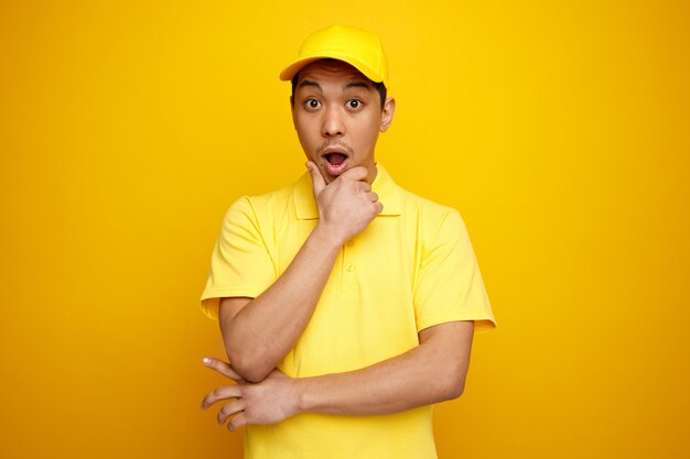 Surprised young delivery man wearing cap and uniform keeping hand on chin 