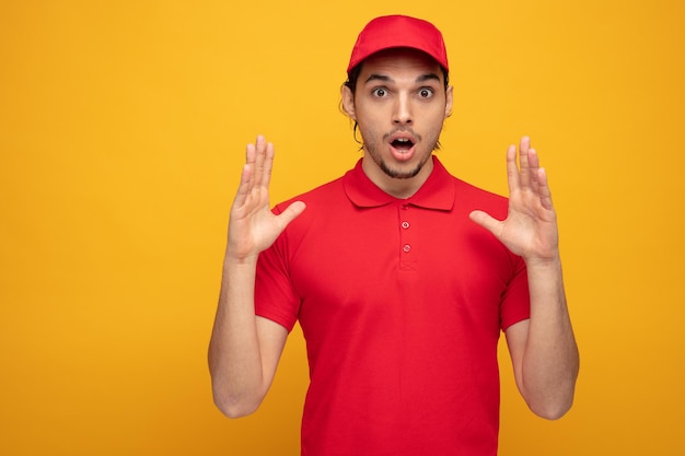 Surprised young delivery man wearing uniform and cap looking at camera while keeping hands in air isolated on yellow background
