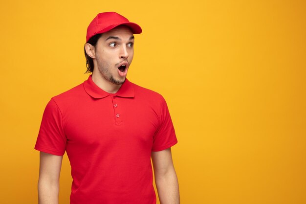surprised young delivery man wearing uniform and cap looking at side isolated on yellow background with copy space
