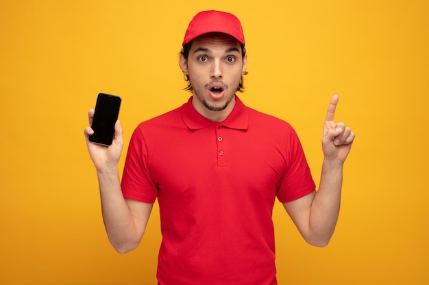 노란색 배경에 격리된 카메라를 쳐다보고 있는 휴대전화를 보여주는 유니폼과 모자를 쓴 젊은 배달원