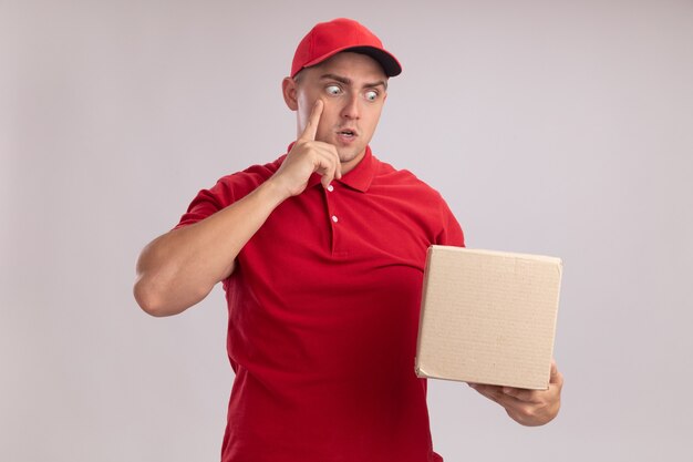 Surprised young delivery man wearing uniform with cap holding and looking at box putting finger on temple isolated on white wall
