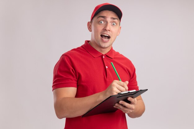 Surprised young delivery man wearing uniform with cap writing something in clipboard isolated on white wall