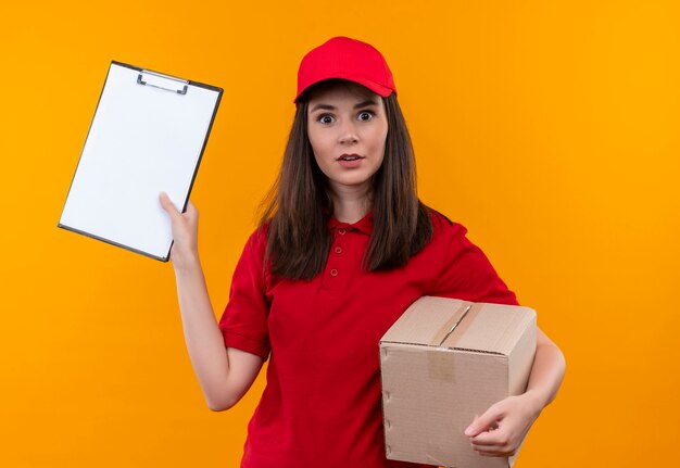 격리 된 노란색 벽에 상자와 클립 보드를 들고 빨간 모자에 빨간 티셔츠를 입고 놀란 젊은 배달 여자