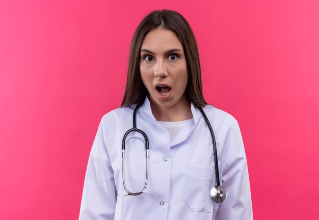 孤立したピンクの壁に聴診器の医療用ガウンを着て驚いた若い医者の女の子